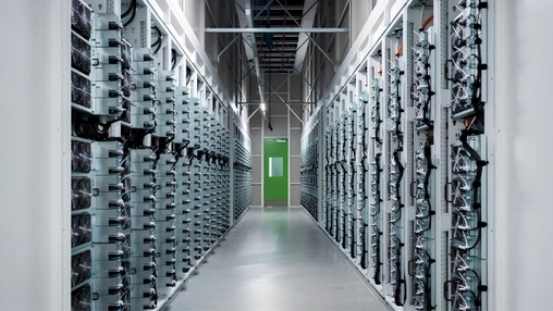 Dense rows of server racks and cooling fans fill a data centre aisle, with a green fire door at the far end