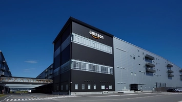 Amazon warehouse with a black front wall showing the Amazon logo and a long grey building beside it